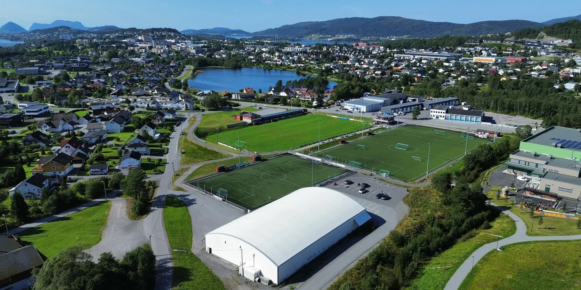 Spjelkavik IL sitt anlegg sett fra lufta, med gressbane, to kunstgressbaner og hall, og Spjelkavik og Ålesund i bakgrunnen.